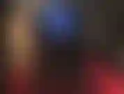 Com leggings azuis metálicas, botas pretas altas e brilhantes e um strapon branco no meio.